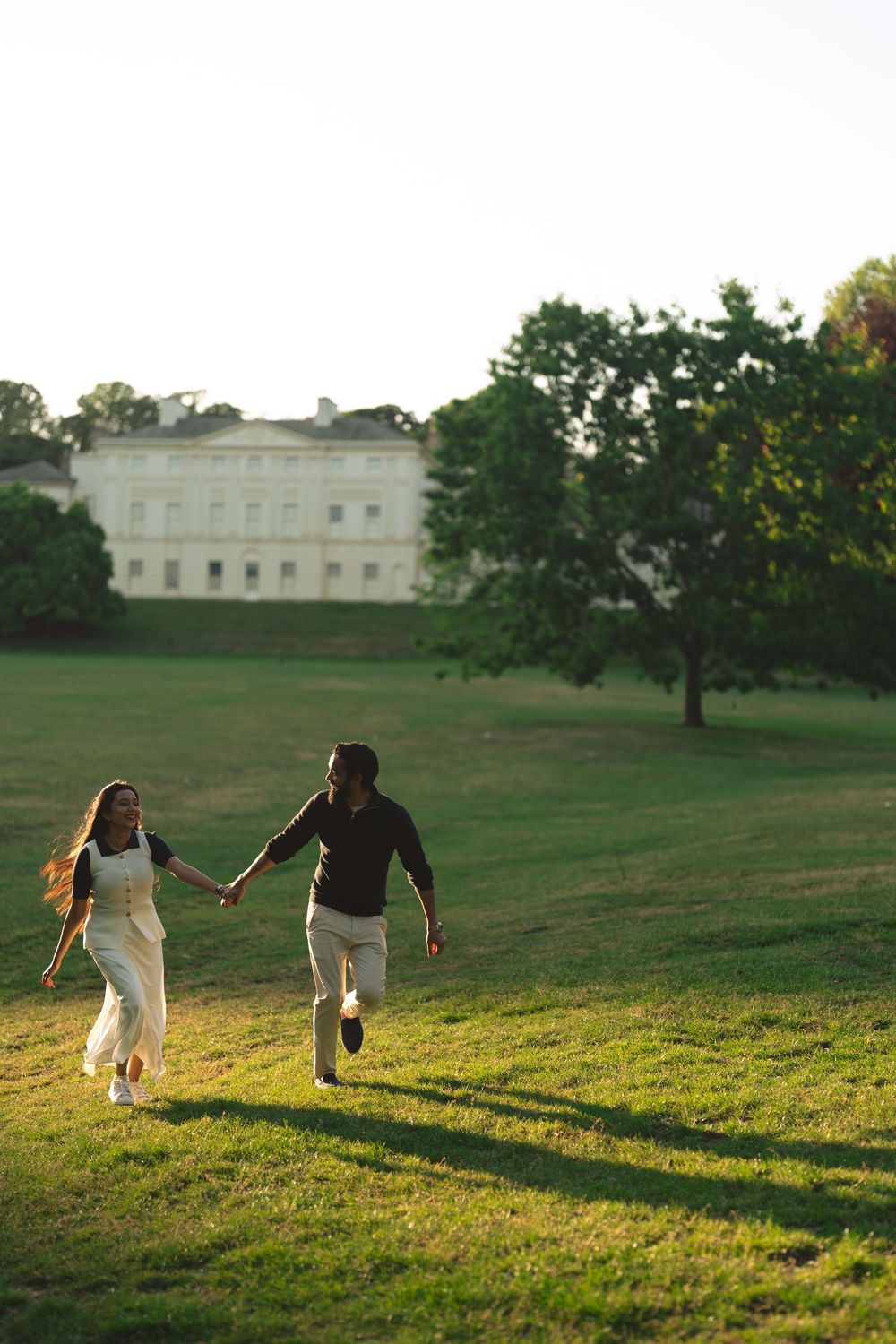 Couple holding hands running through a sunlit green park with a grand white building and trees in the background.