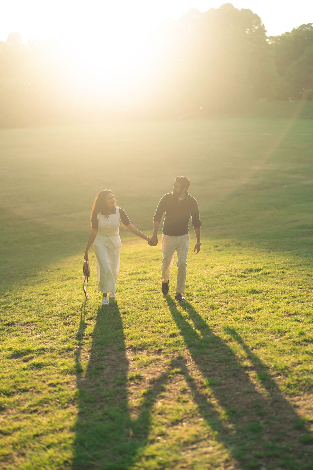 Couple holding hands walking on green grass field bathed in golden sunset light with long shadows.