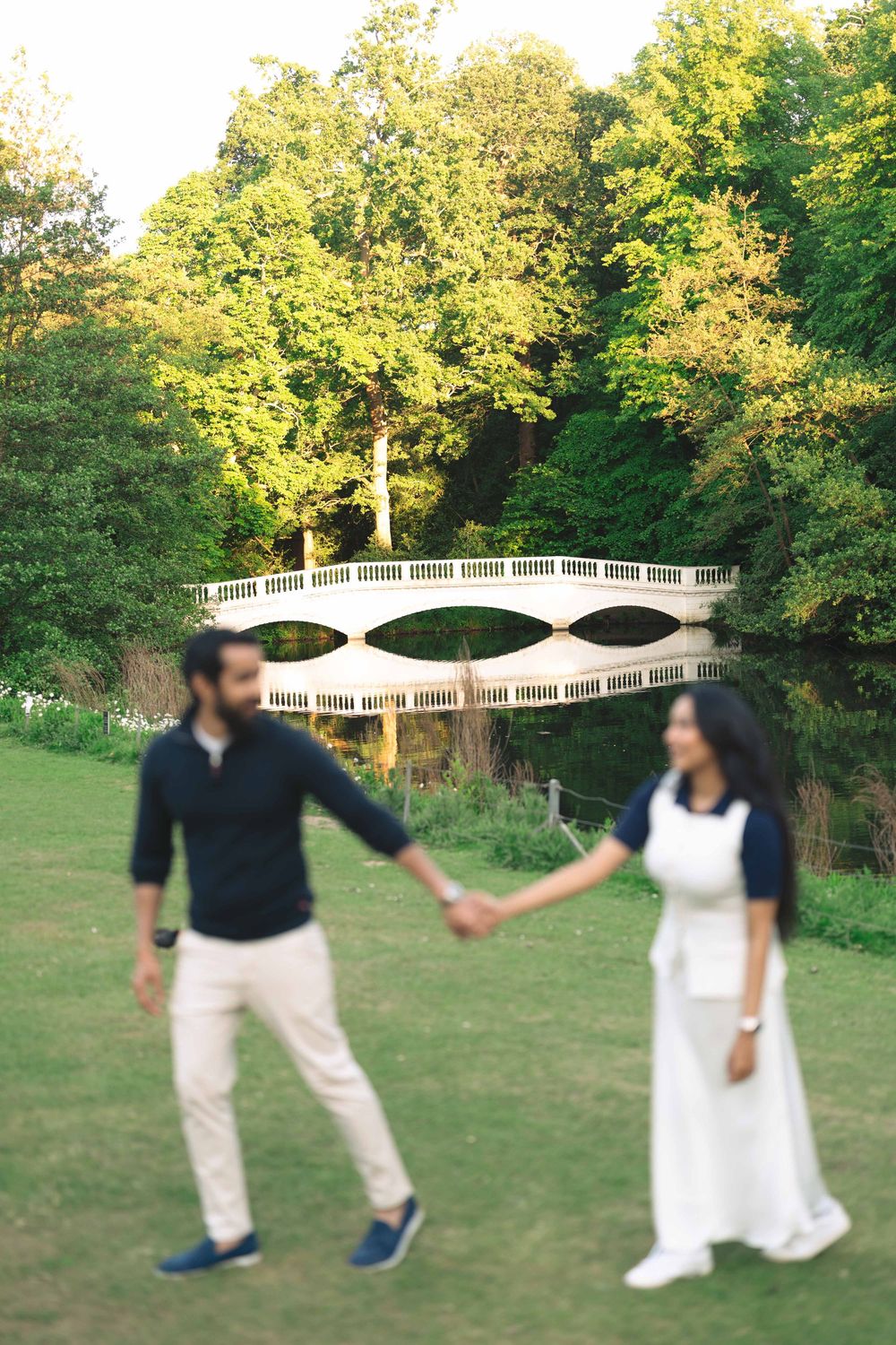 Couple holding hands in a lush green park with a white ornamental bridge and trees in the background.