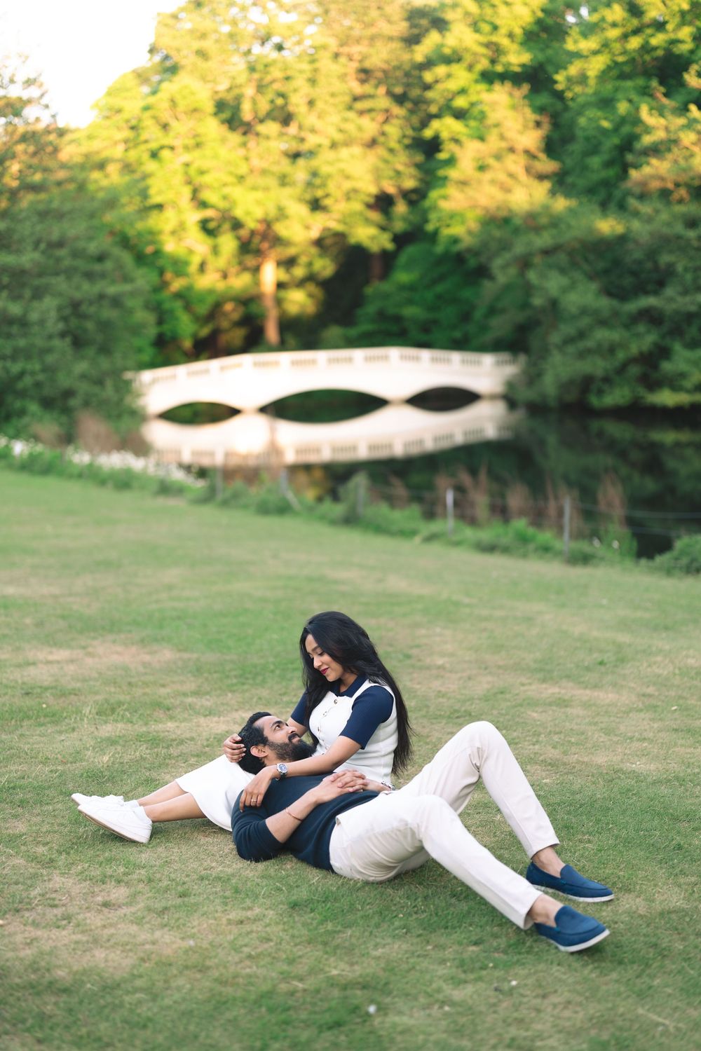 Couple relaxing on green grass in a park, man lying with head on woman's lap near a white bridge.