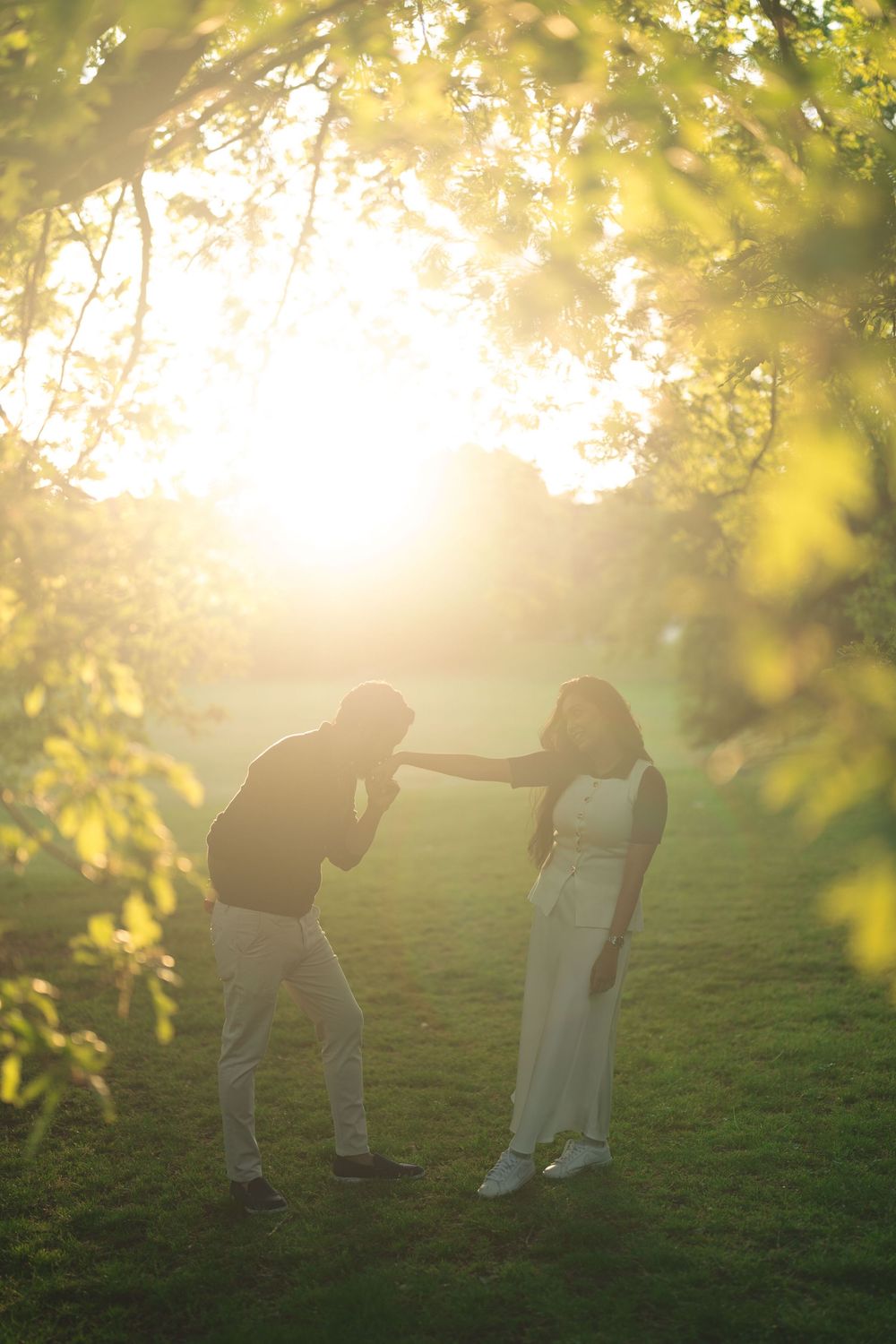 A couple shares a romantic moment in a sunlit park, man bowing to kiss woman's hand amid glowing golden trees.