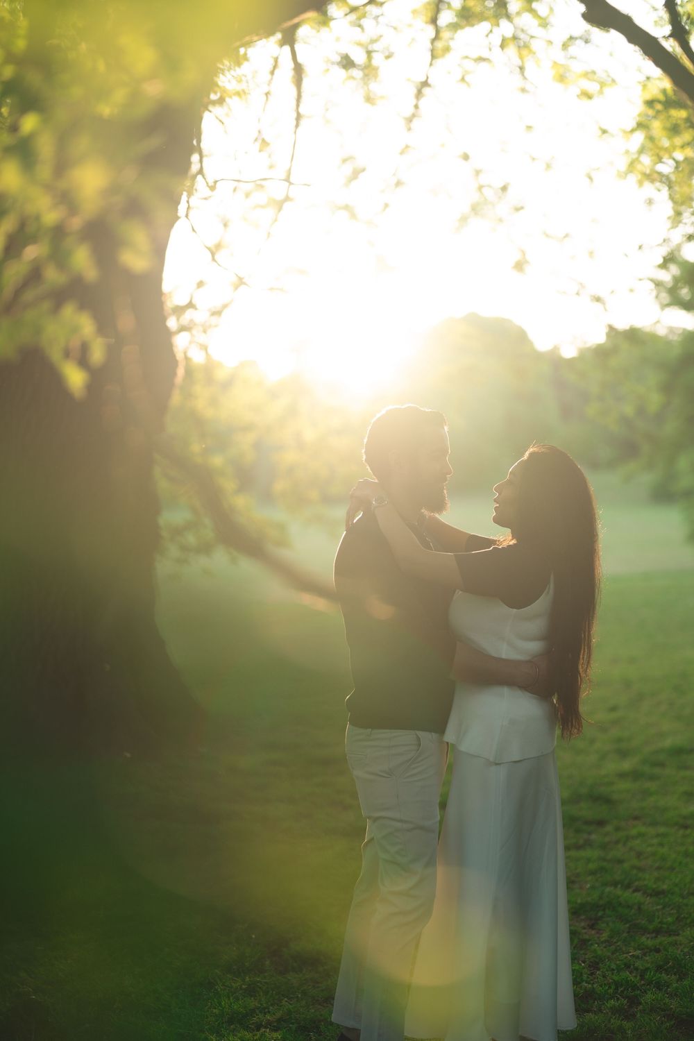 Couple embracing in golden sunset light among lush green trees in a romantic outdoor park setting.