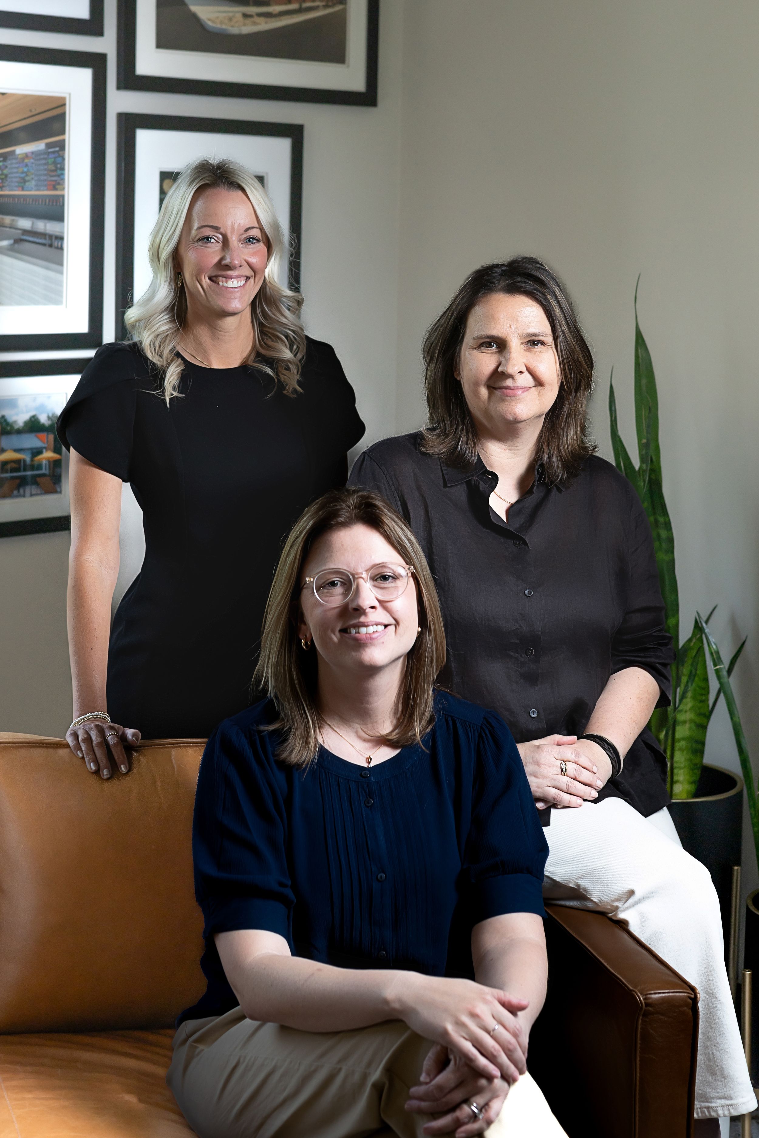 Three professional women pose together in a modern office setting, two standing and one seated on a leather chair.