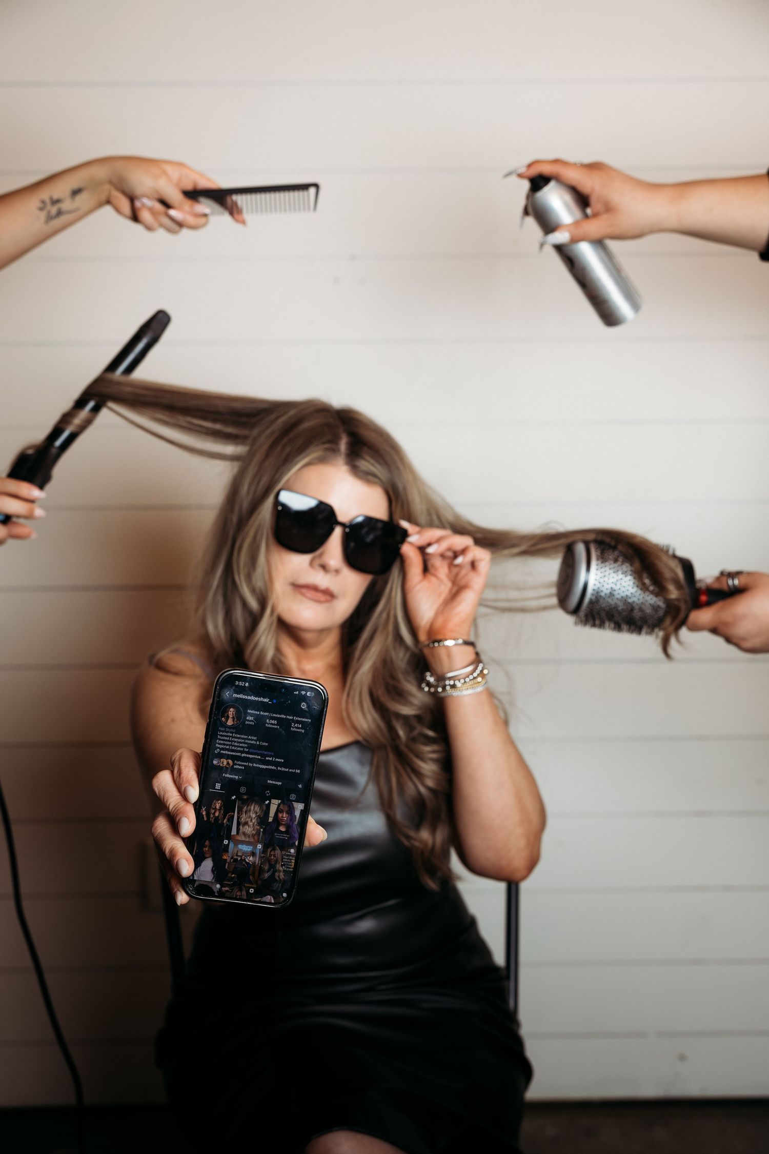 Woman in sunglasses holds phone showing social media while stylists style her hair with tools around her.