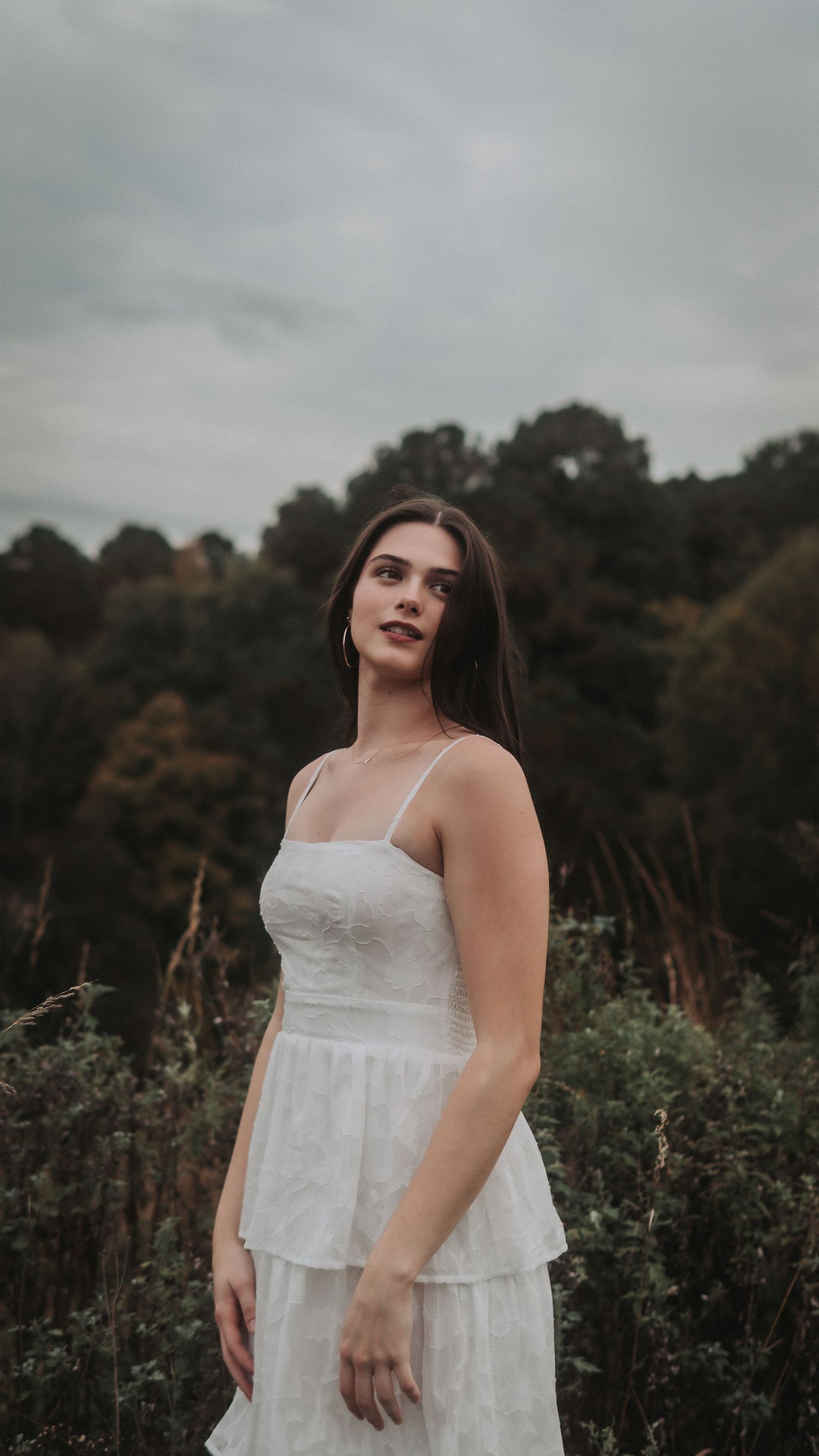 Young woman in white sundress posing outdoors against cloudy sky and green trees.