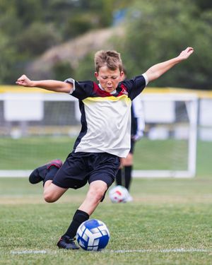Young boy in soccer uniform kicking a blue and white ball on a green grass field during a youth soccer game.