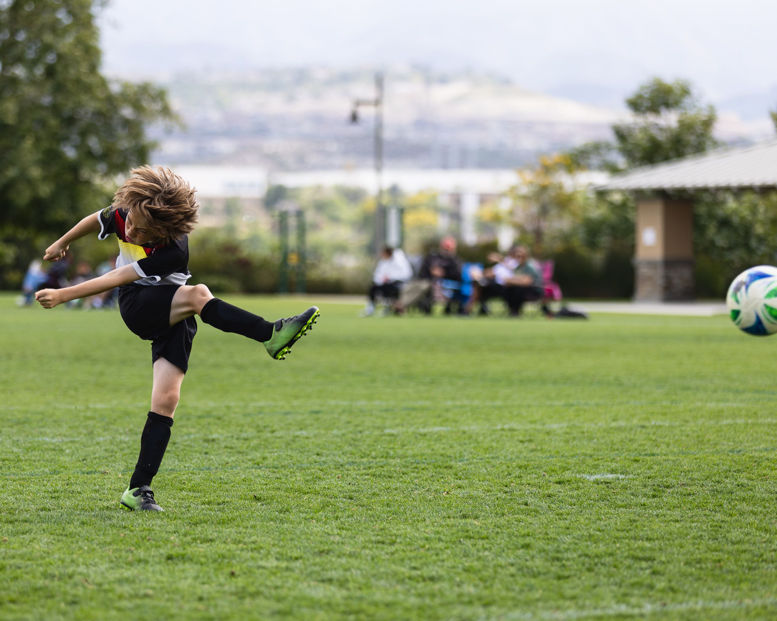 Young soccer player kicking ball on green grass field during youth football match outdoors.