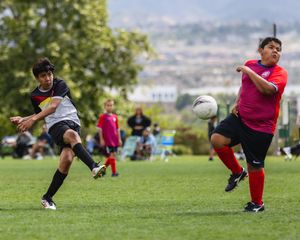 Two youth soccer players compete on a green field, one kicking the ball during an outdoor match.