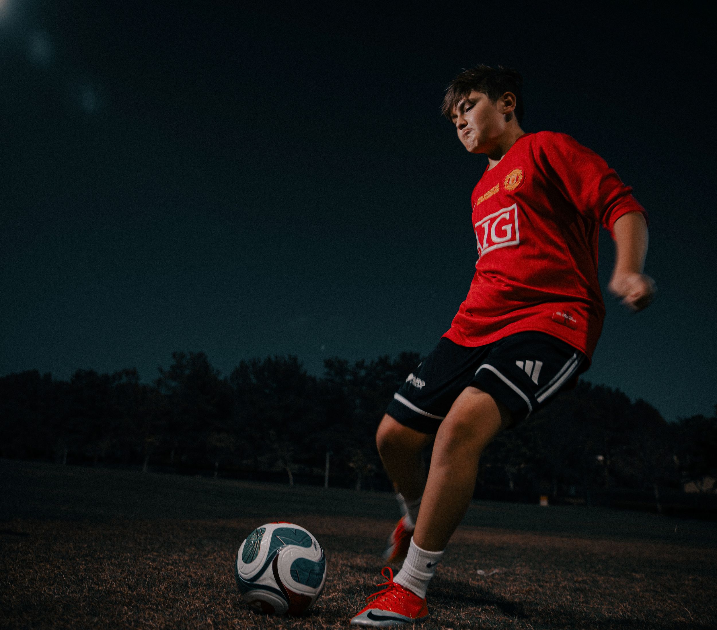 A young boy in a red Manchester United jersey practices soccer skills, kicking a ball on an outdoor court at night.