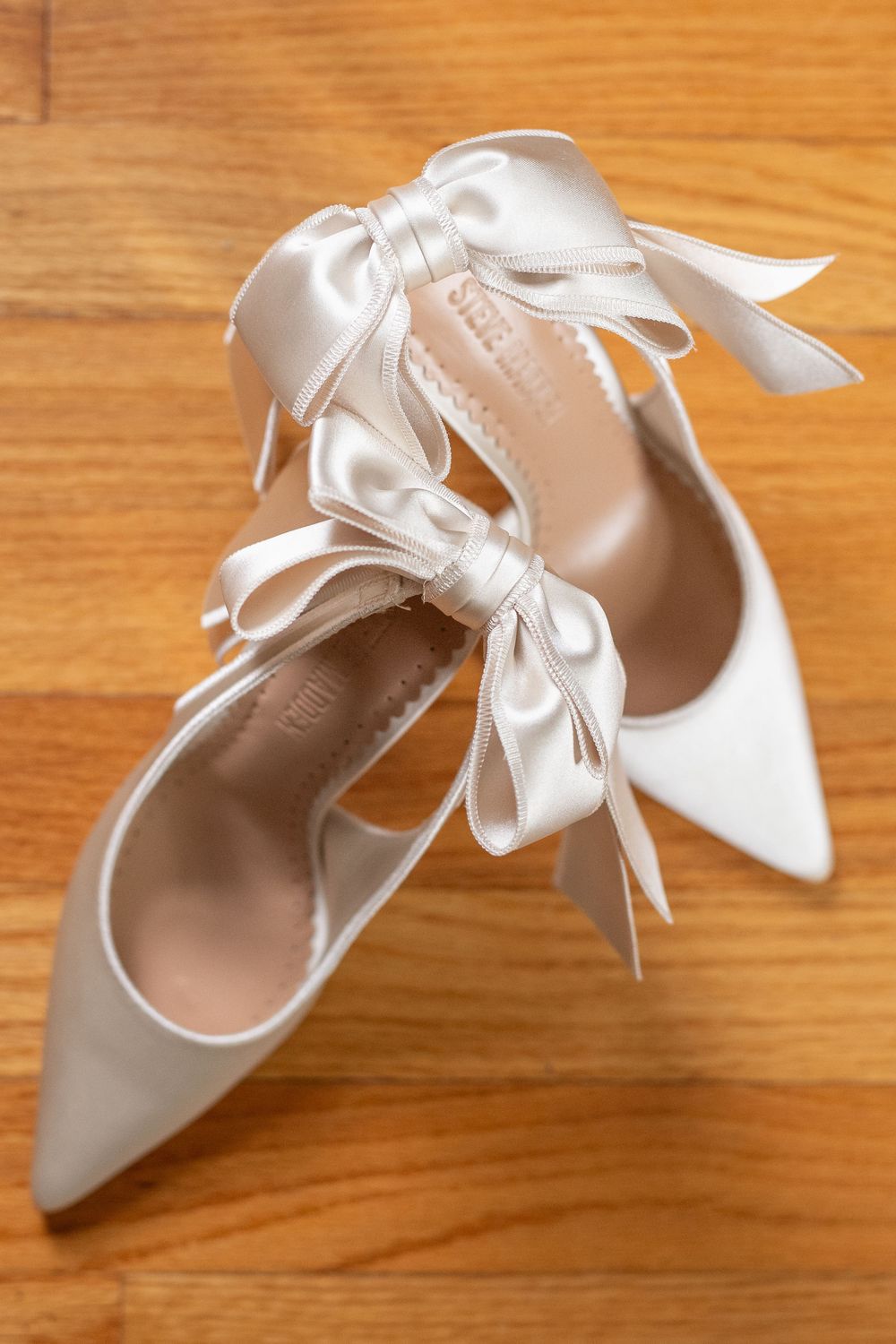 White satin bridal shoes with ribbon bows displayed on wooden floor.