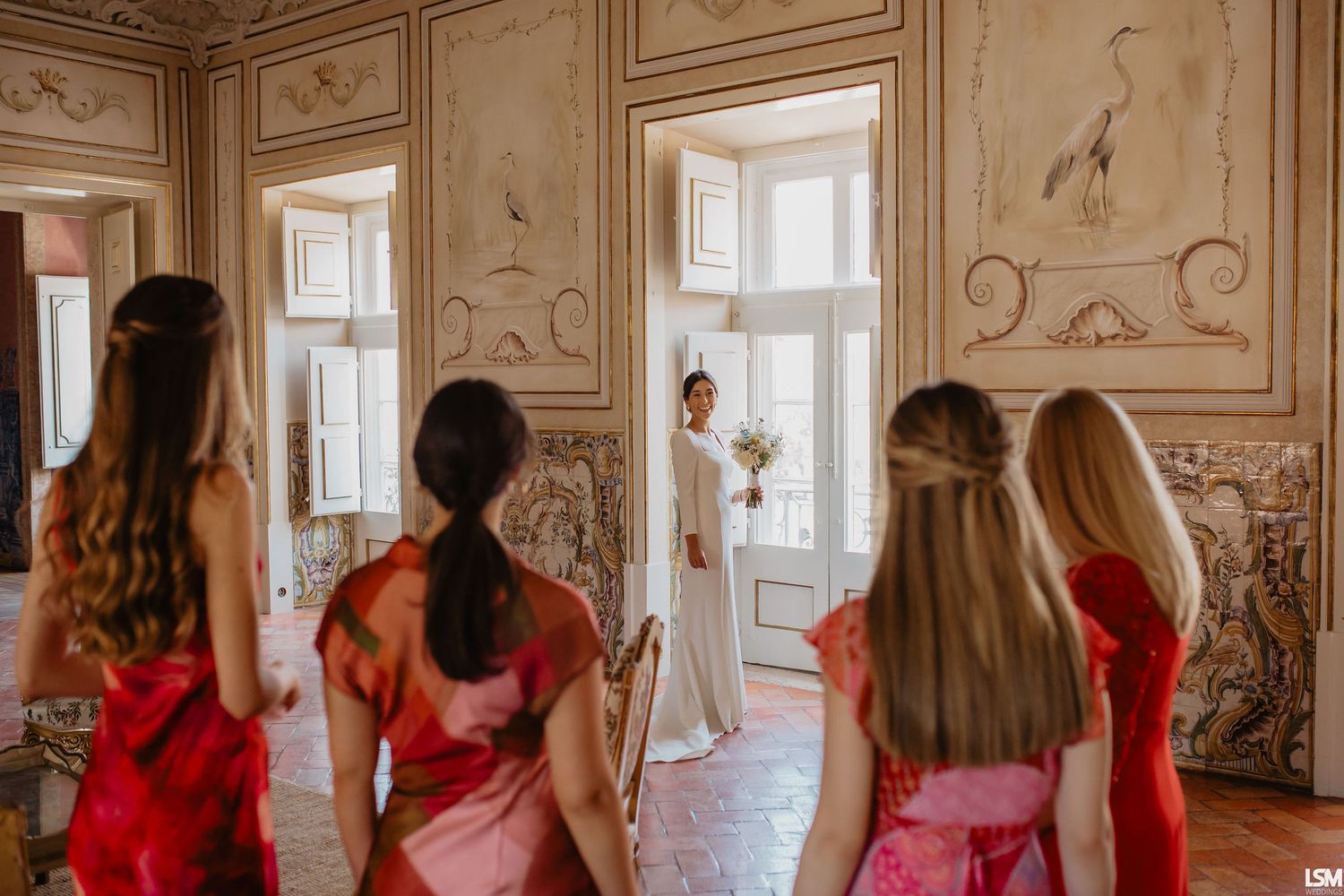 Bridesmaids in red dresses face a bride in white gown standing in an ornate vintage room with decorative walls.