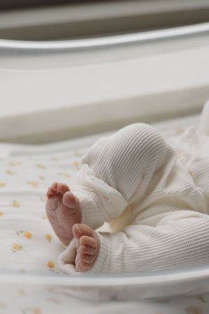 Close-up of tiny newborn baby feet in white onesie lying in hospital bassinet.