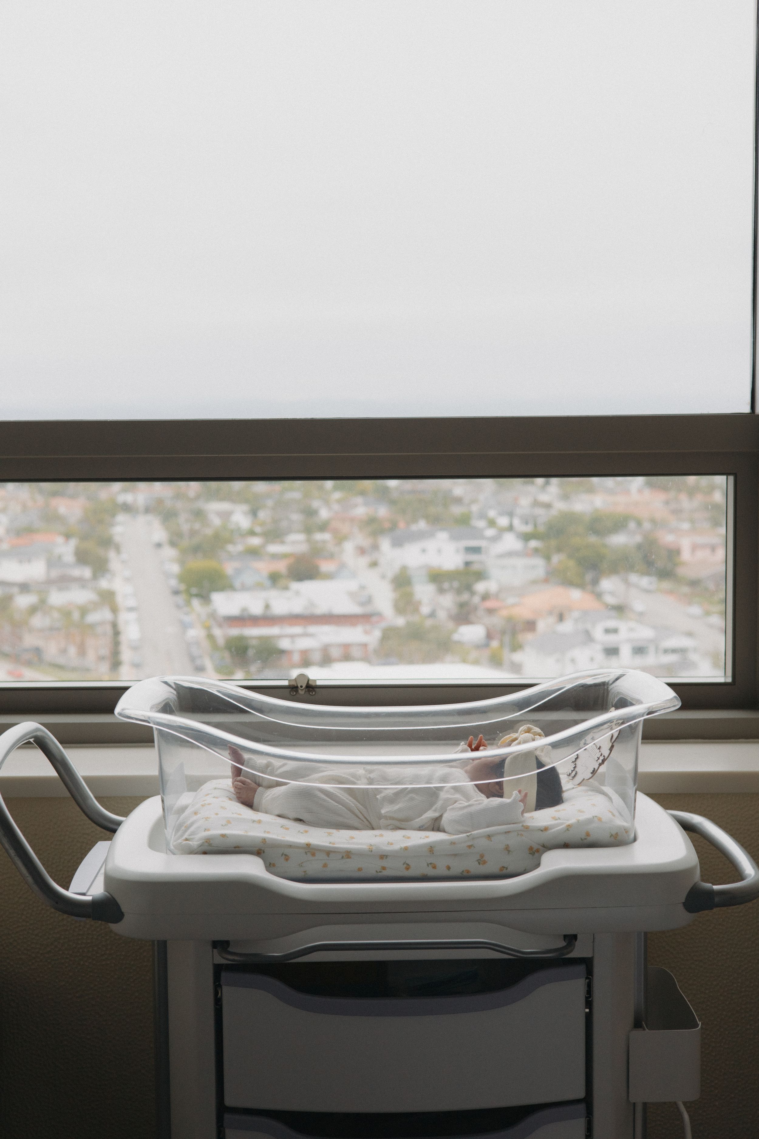 Newborn baby sleeping peacefully in clear hospital bassinet near large window with city view.