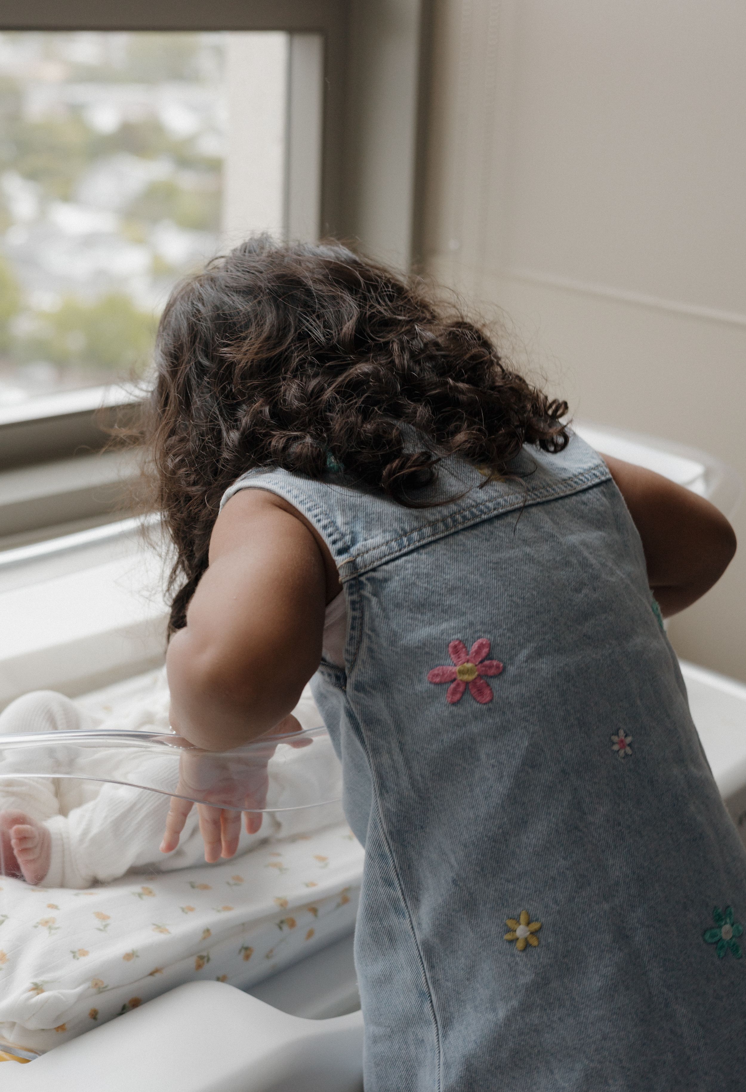 Curly-haired toddler girl in denim dress with floral embroidery leaning over newborn baby bassinet by hospital window.