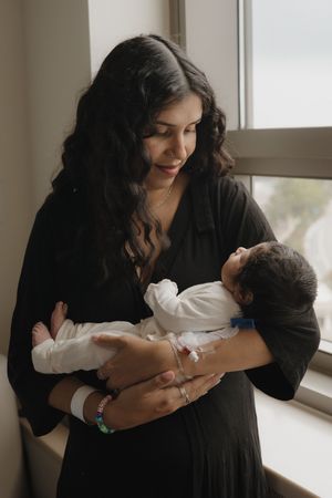 New mother with long dark hair in black dress tenderly holding newborn baby by hospital window.