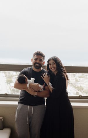 Happy parents smiling and holding newborn baby together in front of large hospital room window.