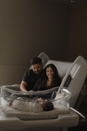 Smiling couple looking lovingly at newborn baby in clear hospital bassinet in dimly lit room.
