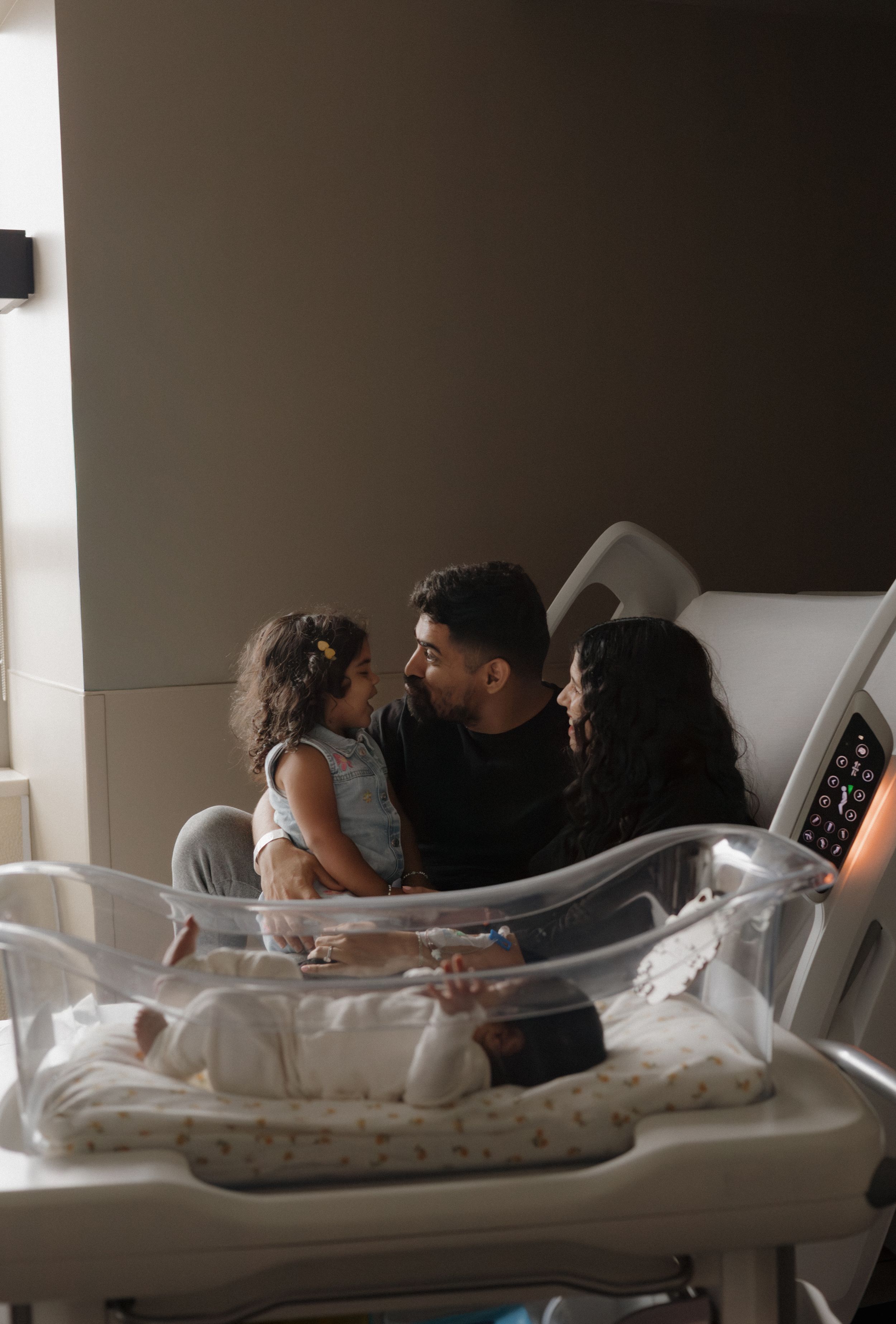 Family of three with toddler and newborn baby in hospital bassinet sharing a tender moment together.