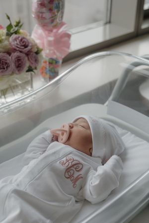 Sleeping newborn baby in white monogrammed hospital outfit with pacifier resting in a clear bassinet beside pink roses.