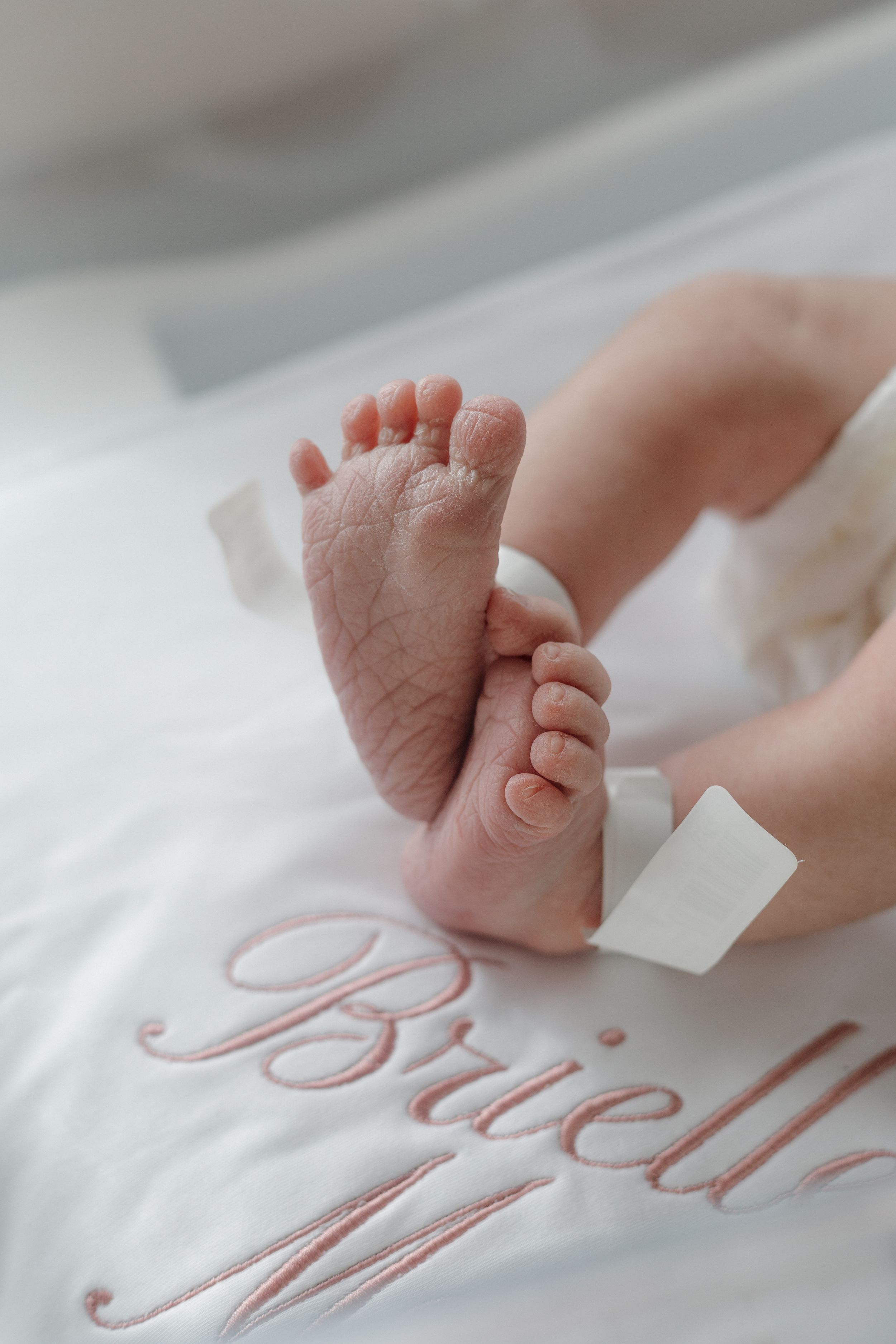 Close-up of newborn baby feet resting on a white blanket embroidered with the name Brielle in pink script lettering.