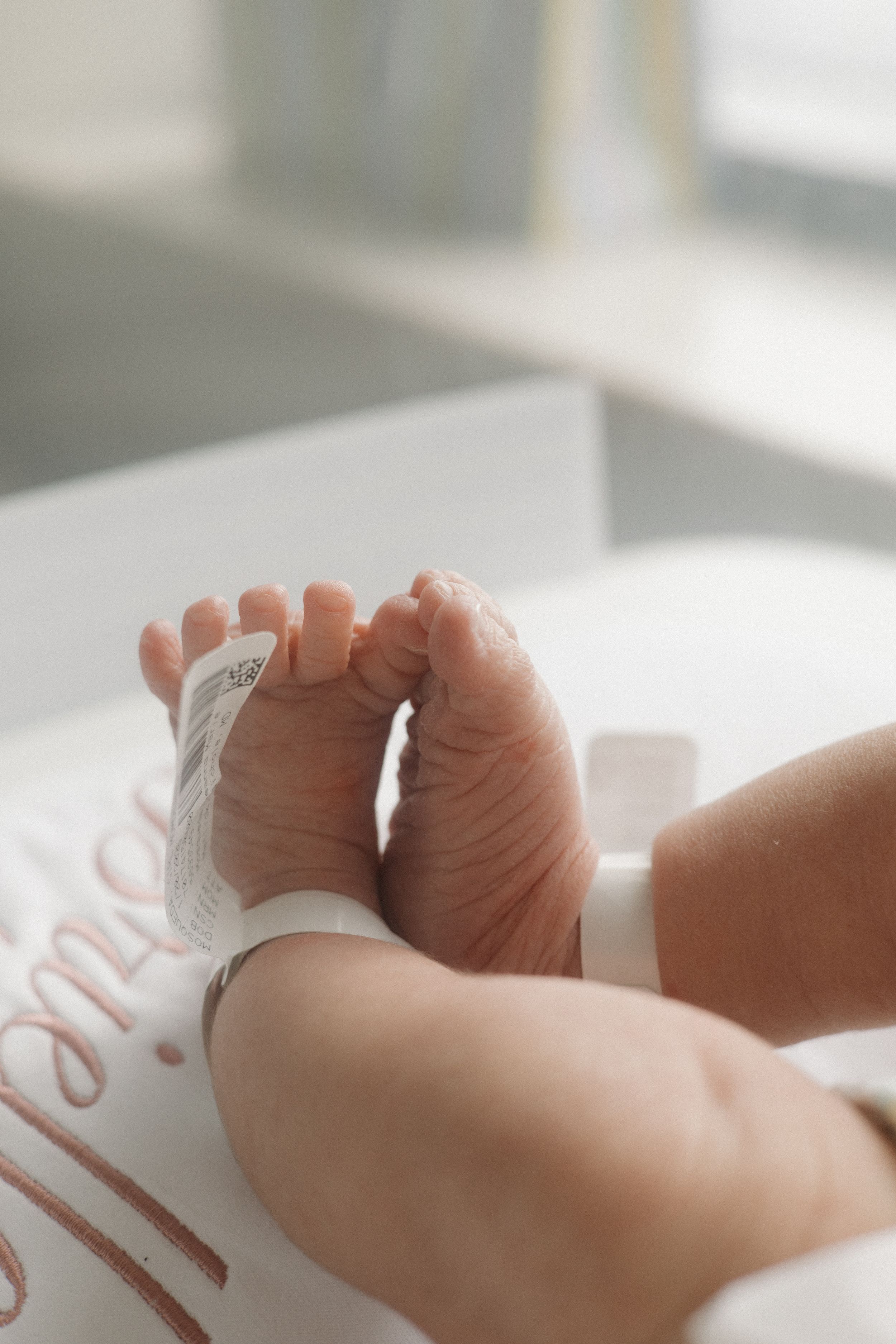 Tiny newborn baby feet with hospital ID bracelet resting on a white personalized name blanket in soft natural light.