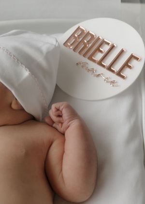 Sleeping newborn baby beside a white and rose gold acrylic name sign reading Brielle with birth details inscribed below.
