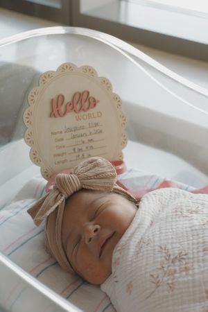 Newborn baby girl swaddled in white with bow headband next to Hello World birth announcement sign.
