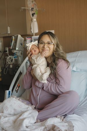 New mother in purple pajamas holding newborn baby girl in hospital room after delivery.