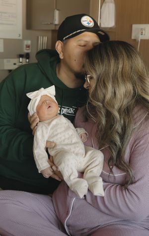 New parents kissing over their newborn baby girl dressed in white lace outfit in hospital.