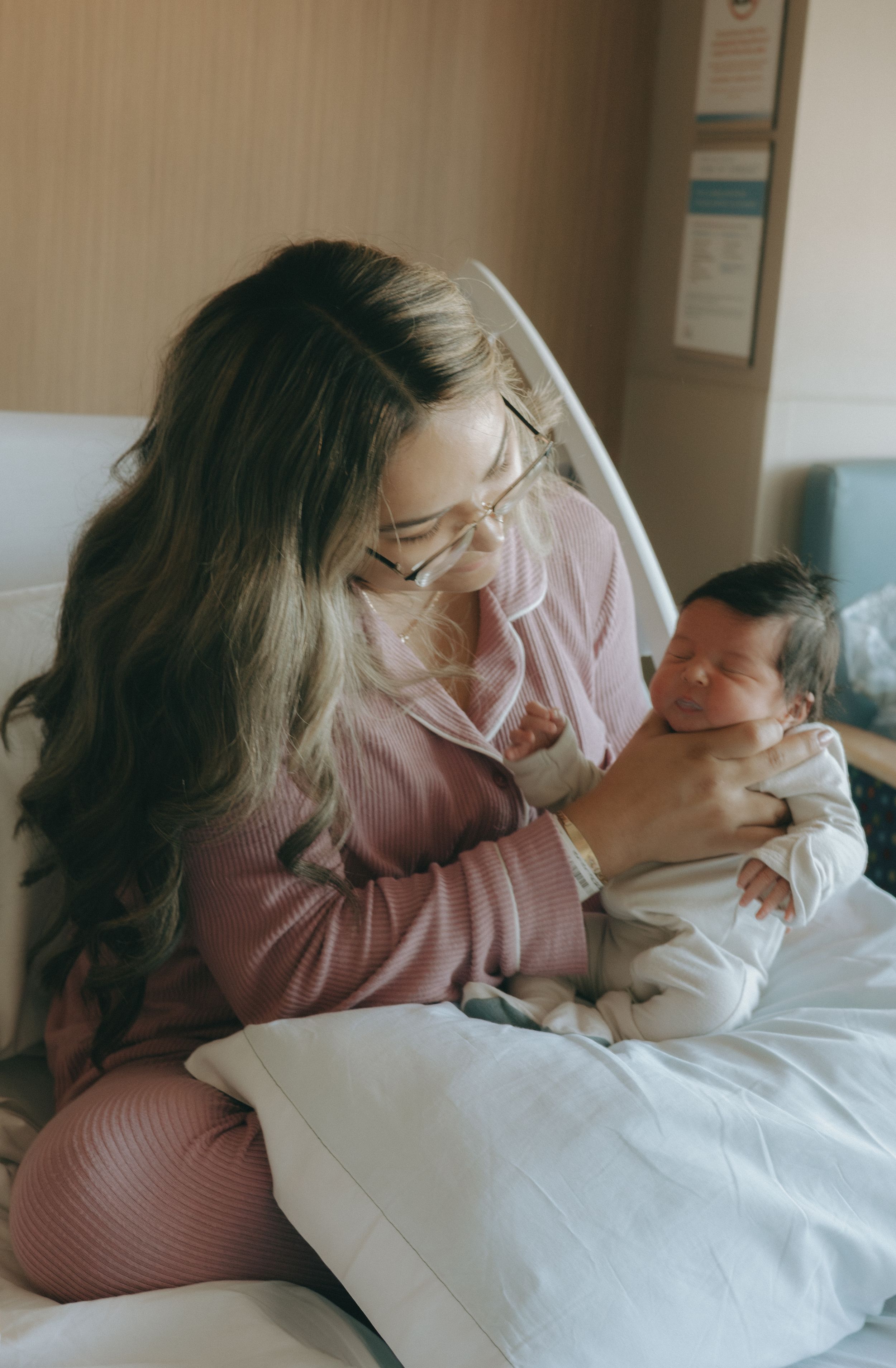 New mother with glasses tenderly holds and gazes at her newborn baby in hospital bed.