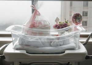 Clear hospital newborn bassinet decorated with pink gift wrap, floral arrangements, and baby gifts by a window.
