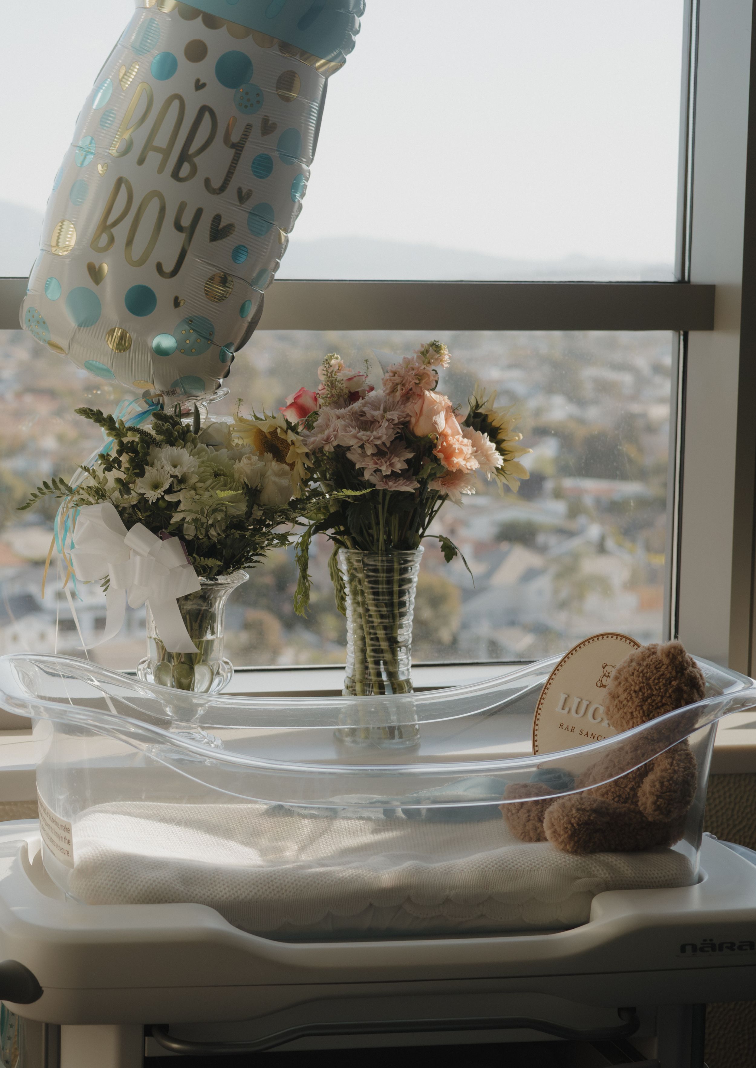 Hospital newborn bassinet by a window decorated with a Baby Boy balloon, flower bouquets, and a teddy bear.