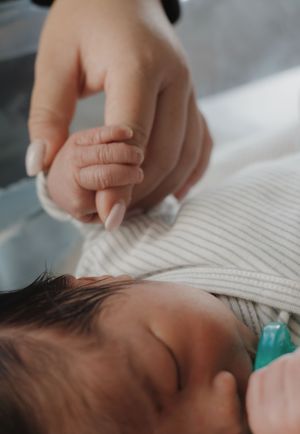 Adult finger gently held by sleeping newborn baby with pacifier in striped hospital swaddle.