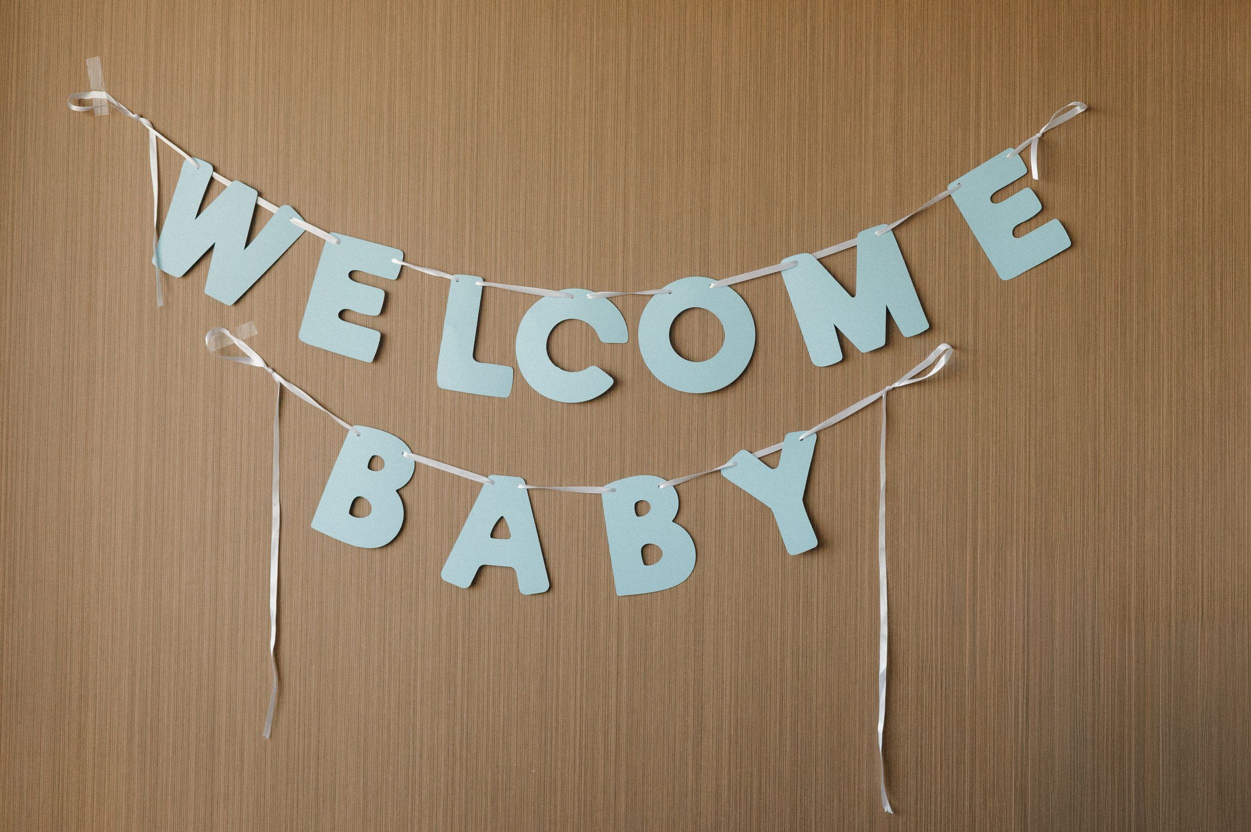 Light blue Welcome Baby letter banner hung on a wooden wall with white ribbon ties for a baby shower decoration.
