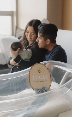 New parents admire their newborn baby beside a clear hospital bassinet with a personalized Lucas Rae Sanchez name sign.