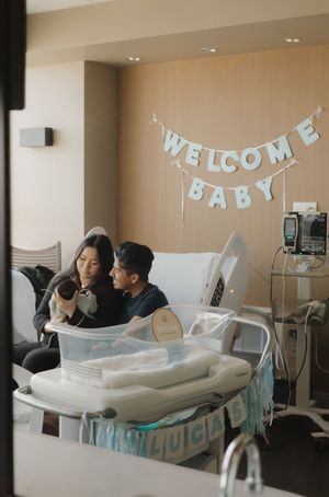 New parents sit together on a hospital bed holding their newborn with a Welcome Baby banner on the wall behind them.
