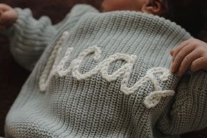 Close-up of a sage green knit baby sweater with the name Lucas embroidered in cream cable knit lettering.