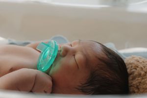 Peaceful newborn baby sleeping with teal pacifier in white hospital bassinet.