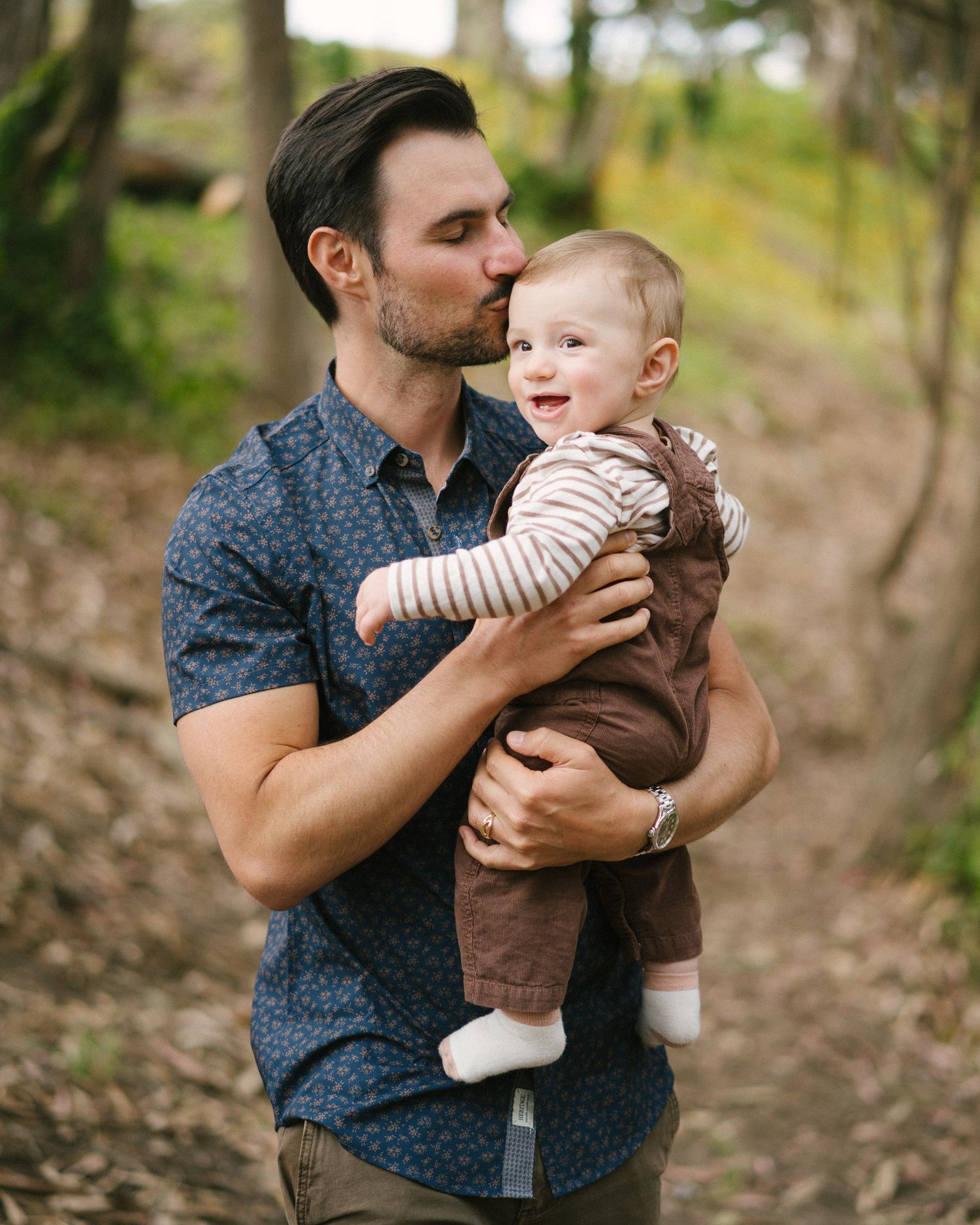 A young man in a blue floral shirt kisses a smiling baby in brown overalls outdoors in a wooded park.