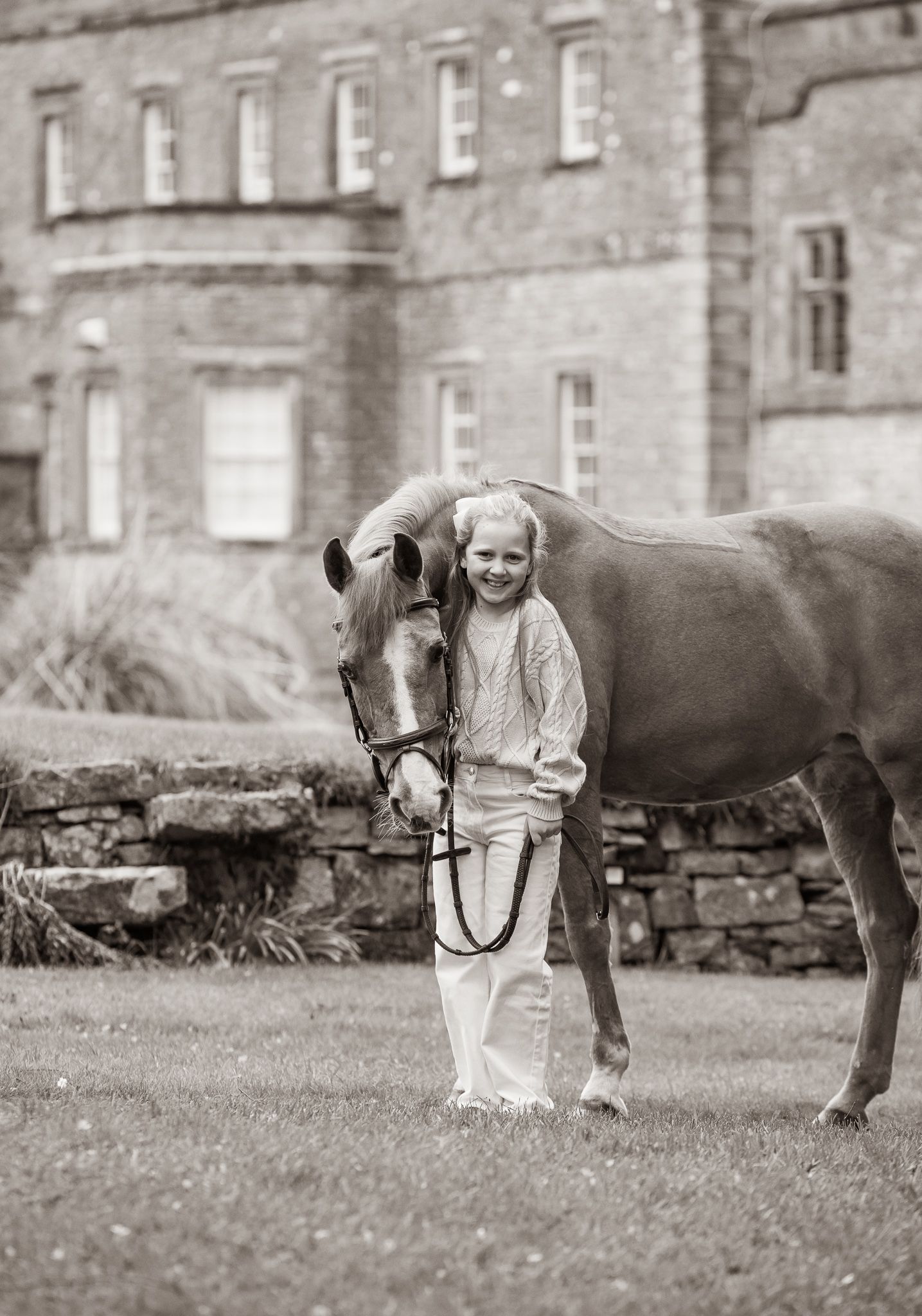Young girl kneeling beside a horse in a black and white countryside photo near a stone building.