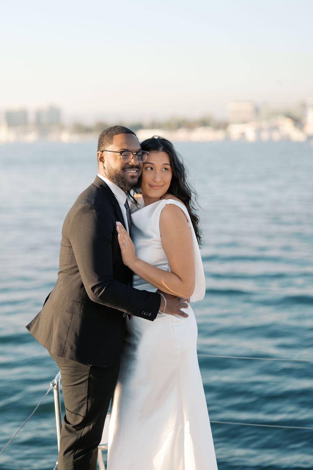 Couple in wedding attire embracing on a boat with calm blue water and city skyline in the background.