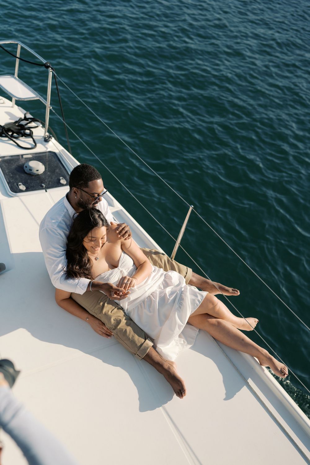 Romantic couple relaxing on sailboat deck, woman in white dress leaning against man over turquoise sea.