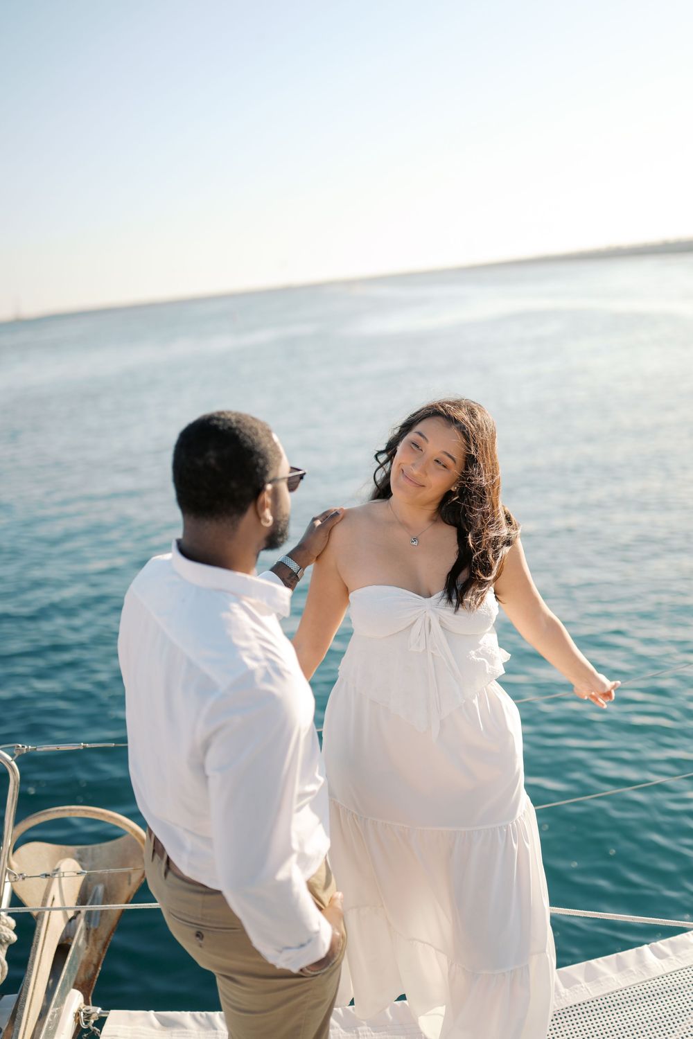 Interracial couple sharing a romantic moment on a yacht, woman in white strapless dress with ocean backdrop.