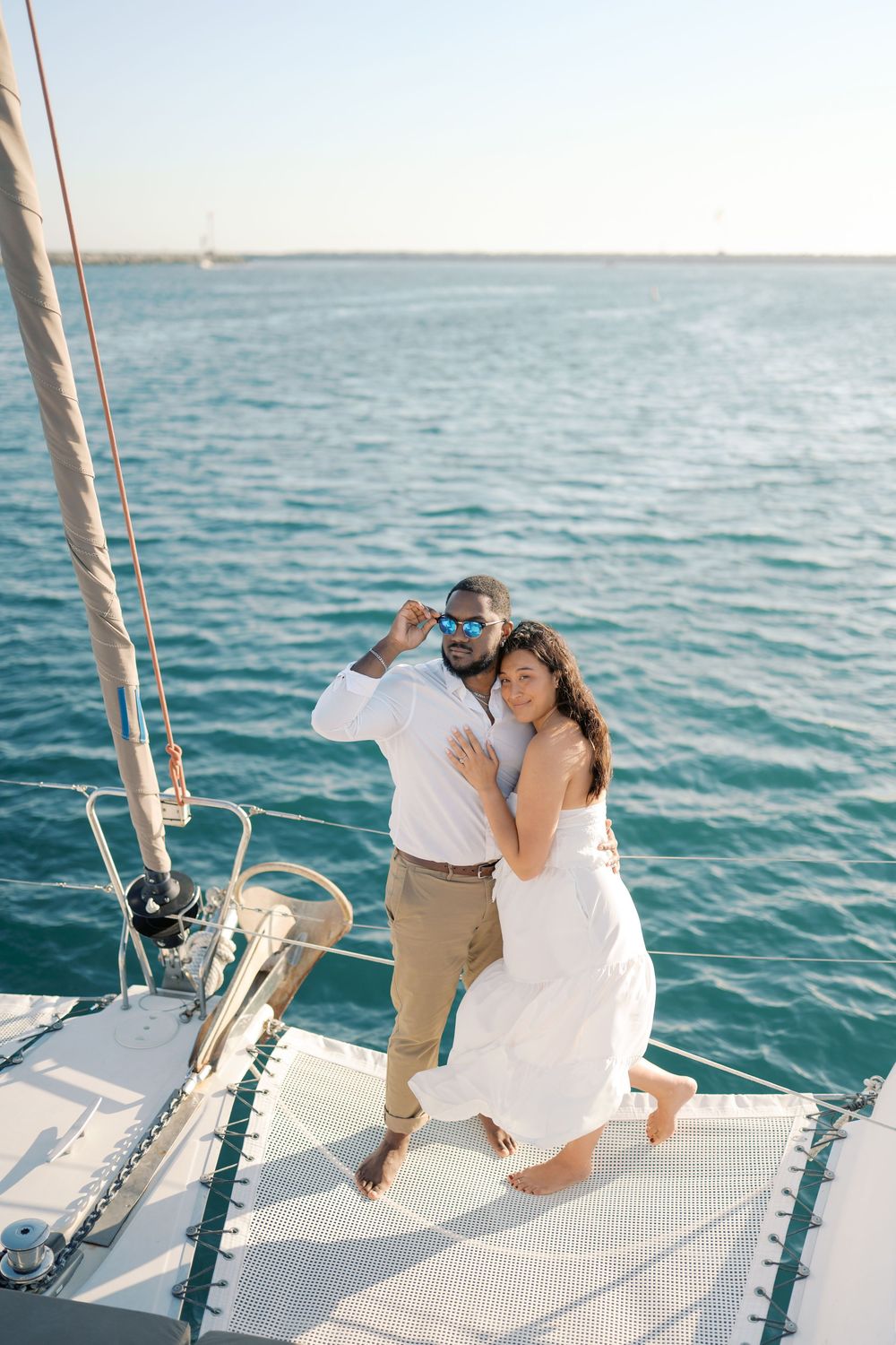 Couple embracing on a white catamaran sailboat deck with turquoise ocean water in the background.