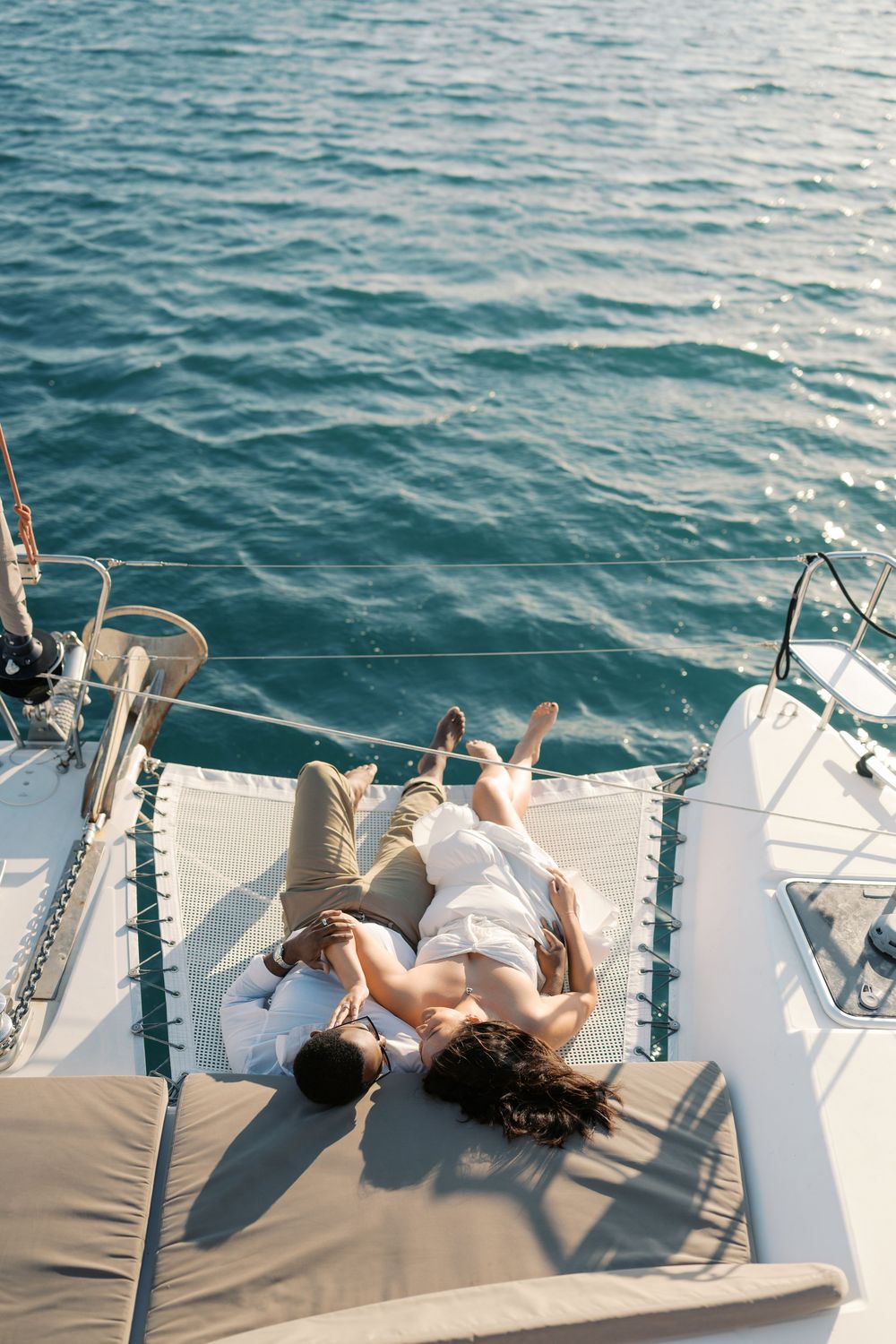 Couple relaxing together on catamaran net over turquoise ocean water on sunny day.
