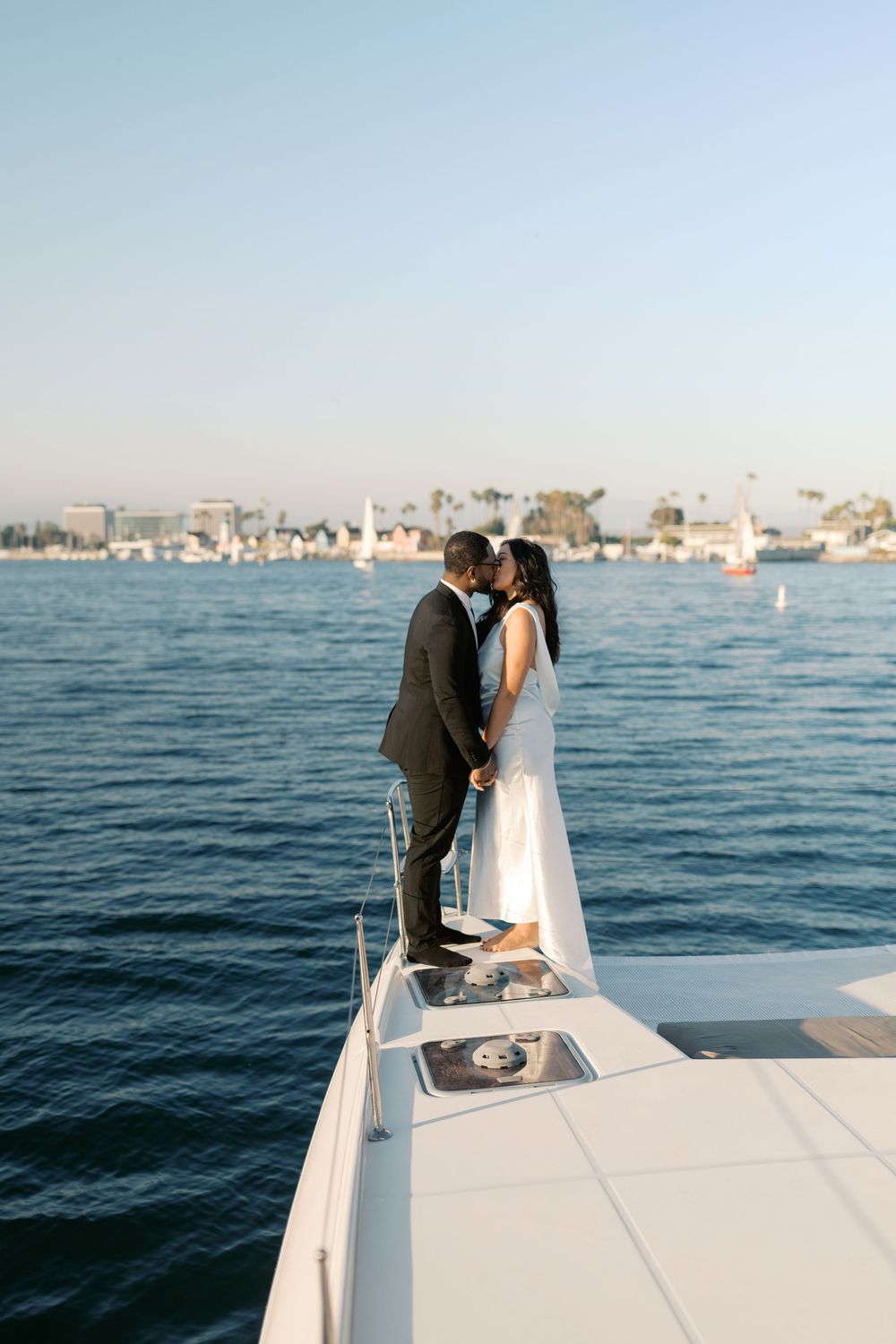Couple sharing a romantic kiss on the bow of a white yacht with a harbor skyline backdrop.