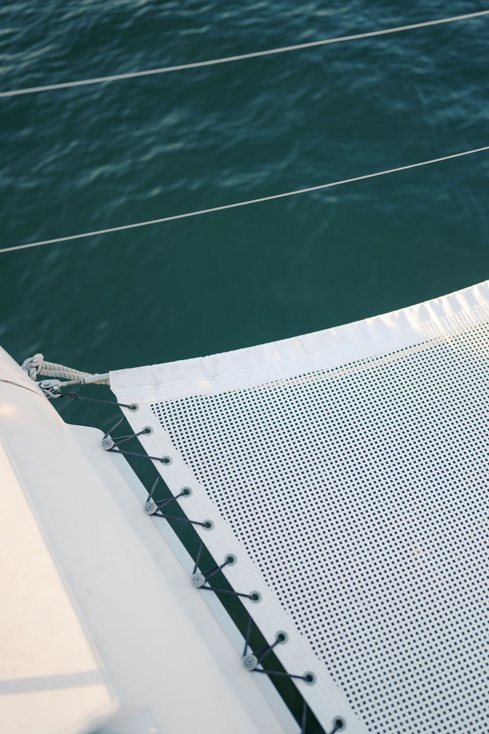 Close-up of white catamaran trampoline mesh netting with rigging lines over turquoise sea water.