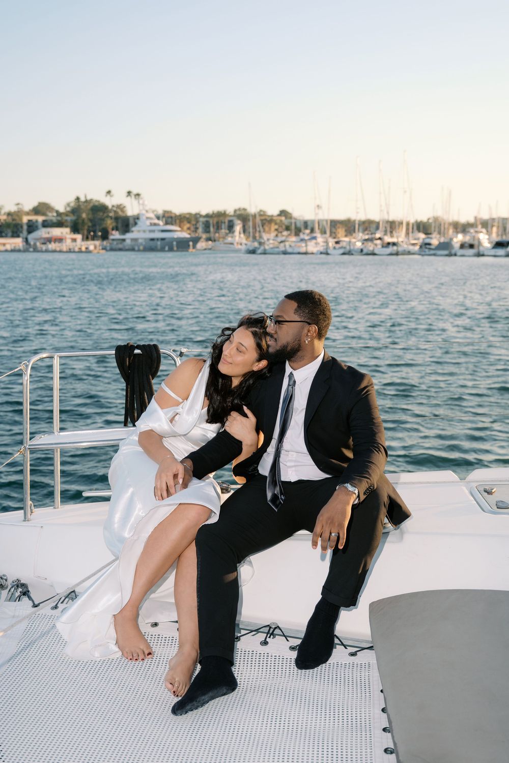 Newlywed couple sitting closely together on a yacht deck with a marina and sailboats behind them.