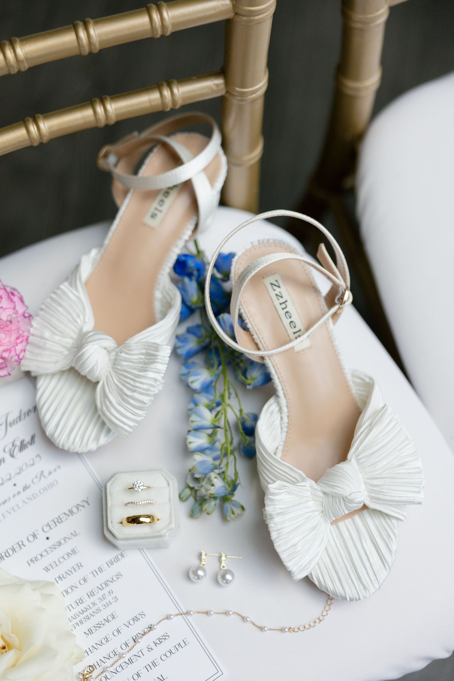 White bridal heels with bow detail displayed alongside pearl earrings, ring box, and floral wedding stationery on chair.