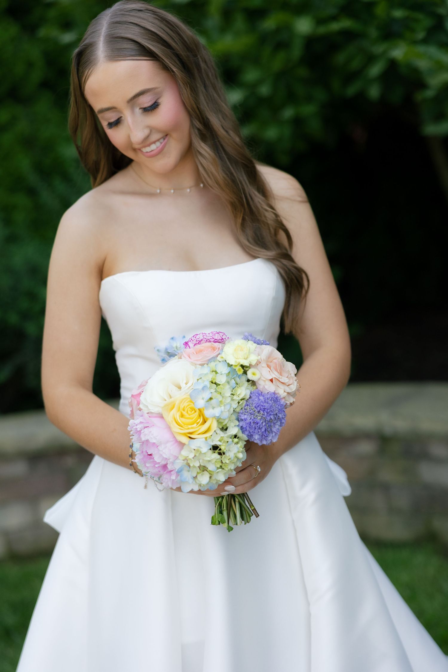 Smiling bride in strapless white gown holding colorful pastel bouquet of roses and hydrangeas in lush garden setting.
